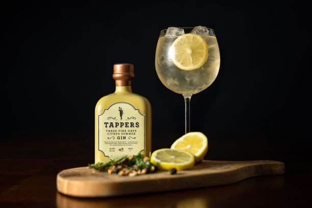 a bottle of gin on a table with a gin and tonic and cut citrus on a wooden board