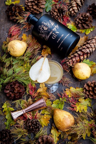 a bottle of brentingby black gin on a table with leaves and a cocktail garnished with pear