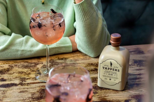 a bottle of gin on a wooden table with a gin and tonic and woman in the background wearing a green jumper