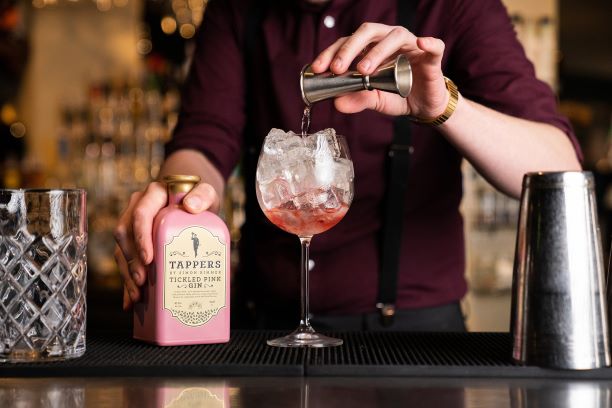 a bottle of pink gin with a gin and tonic on a bar with gin being poured from a jigger